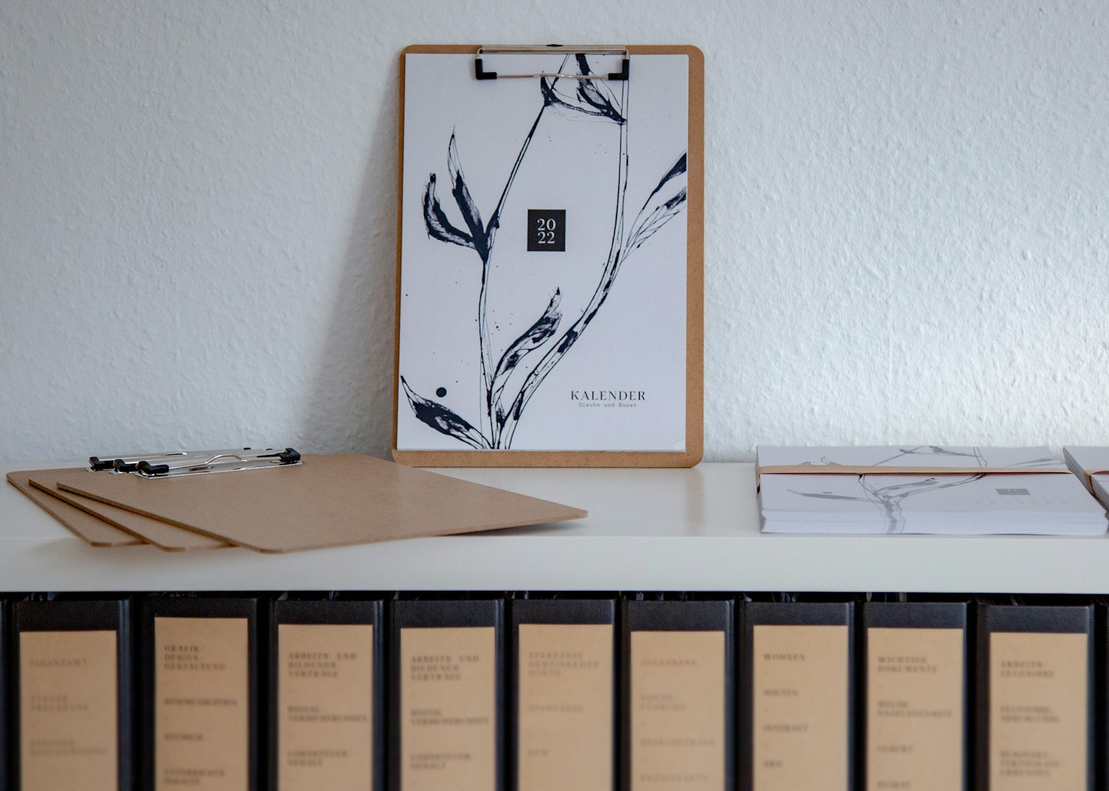 A row of dark binders with labeled spines on a shelf, beneath clipboards and a wall calendar propped against a white wall.