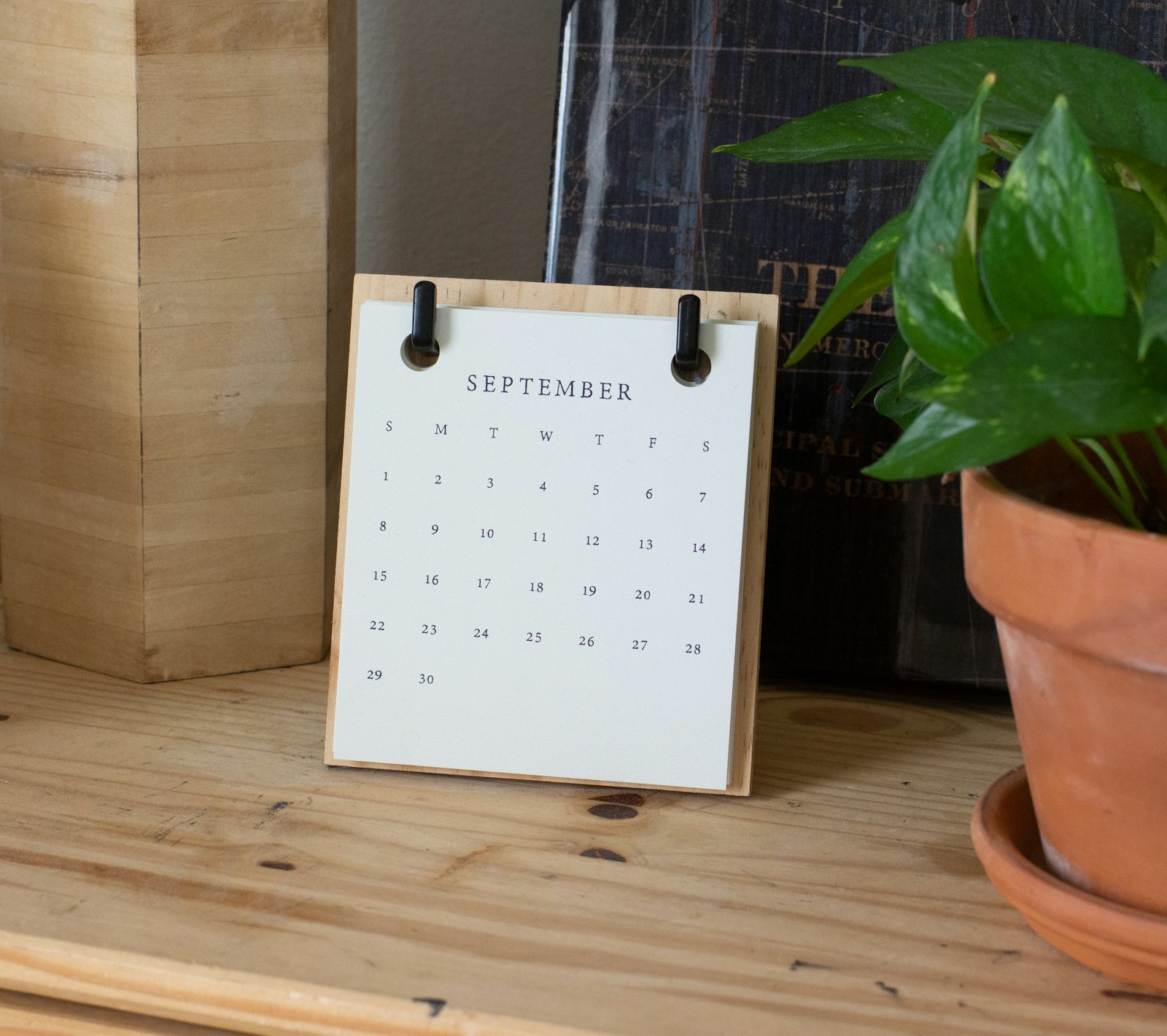 A small desk calendar open to September, on a wooden shelf beside a potted plant.